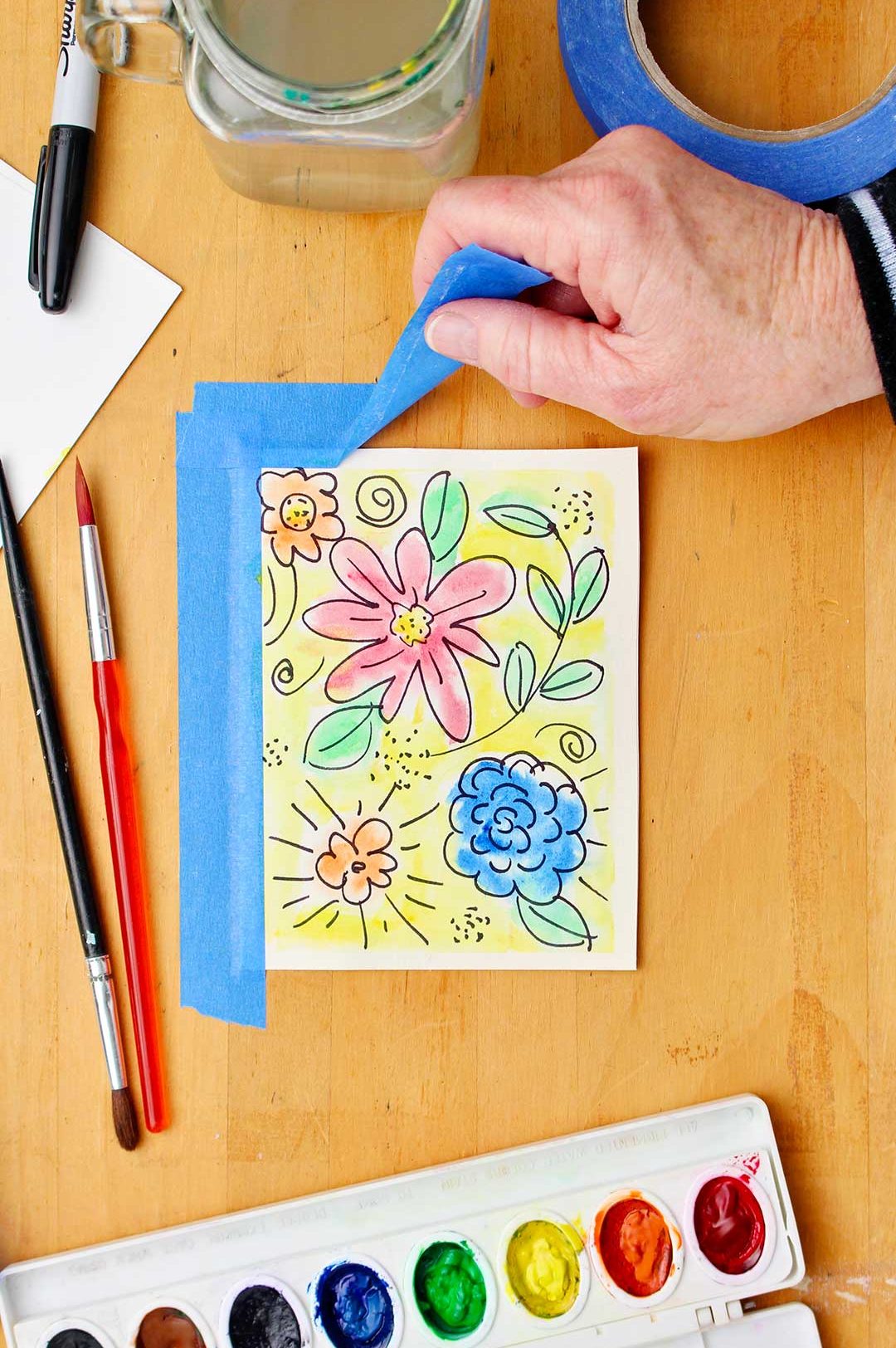 Hand peeling up blue painters tape off wooden table to reveal completed watercolor flower card.