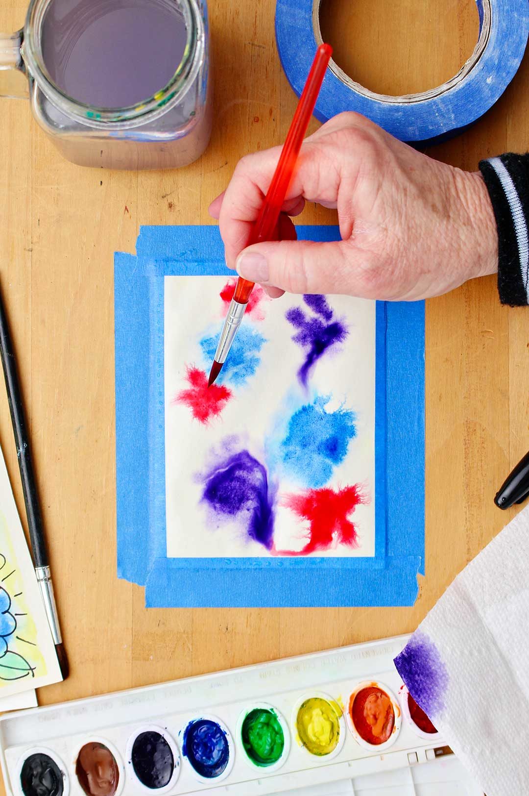 Hand painting abstract splotches of watercolor on a white piece of cardstock taped to the table with watercolor supplies near by.