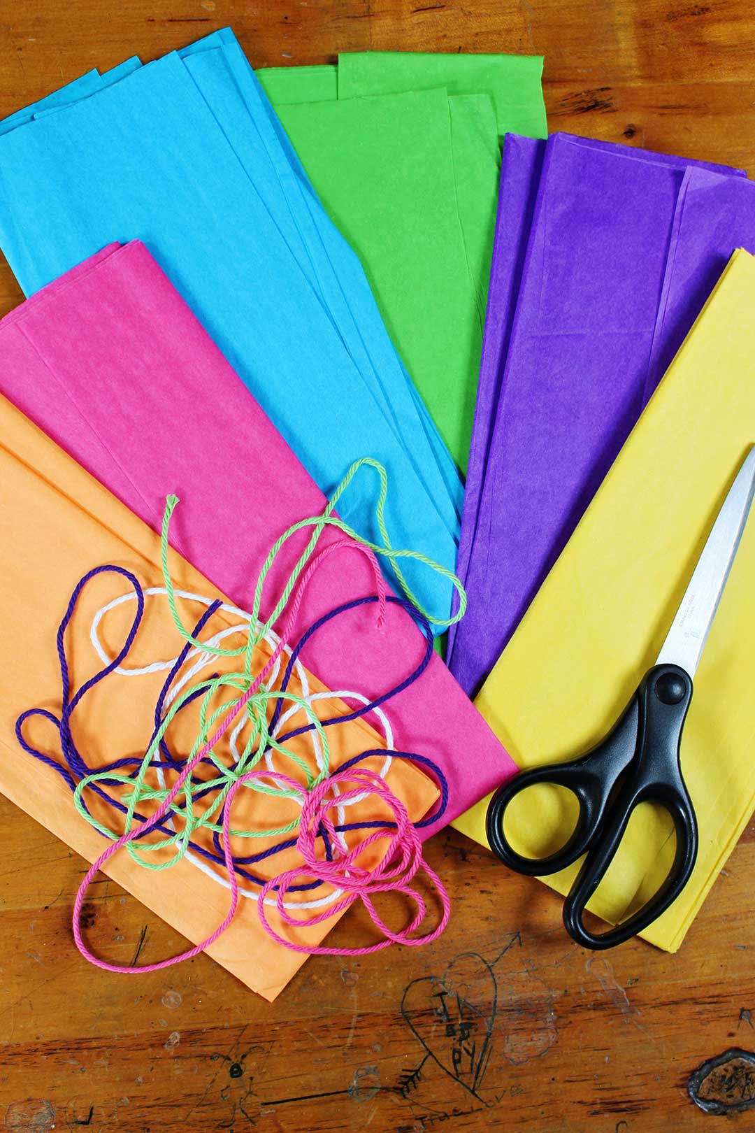 Stacks of orange, pink, blue, green, purple and yellow tissue paper with yarn and scissors resting on them.
