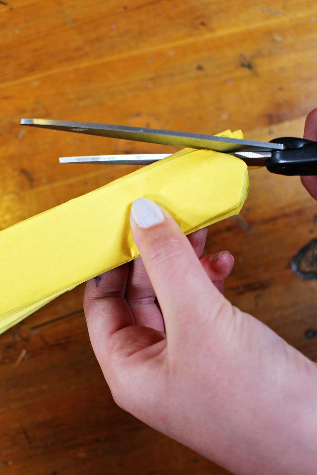 A hand cutting the ends of the folded yellow tissue paper to a rounded edge with scissors.