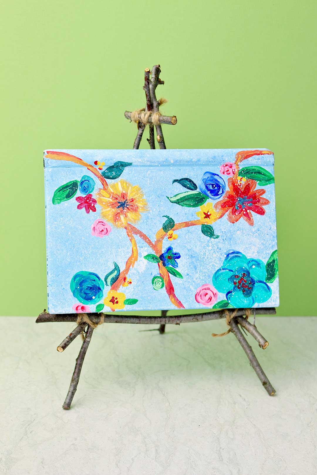 Tabletop easel from twigs holding a painted journal on display against a green background.