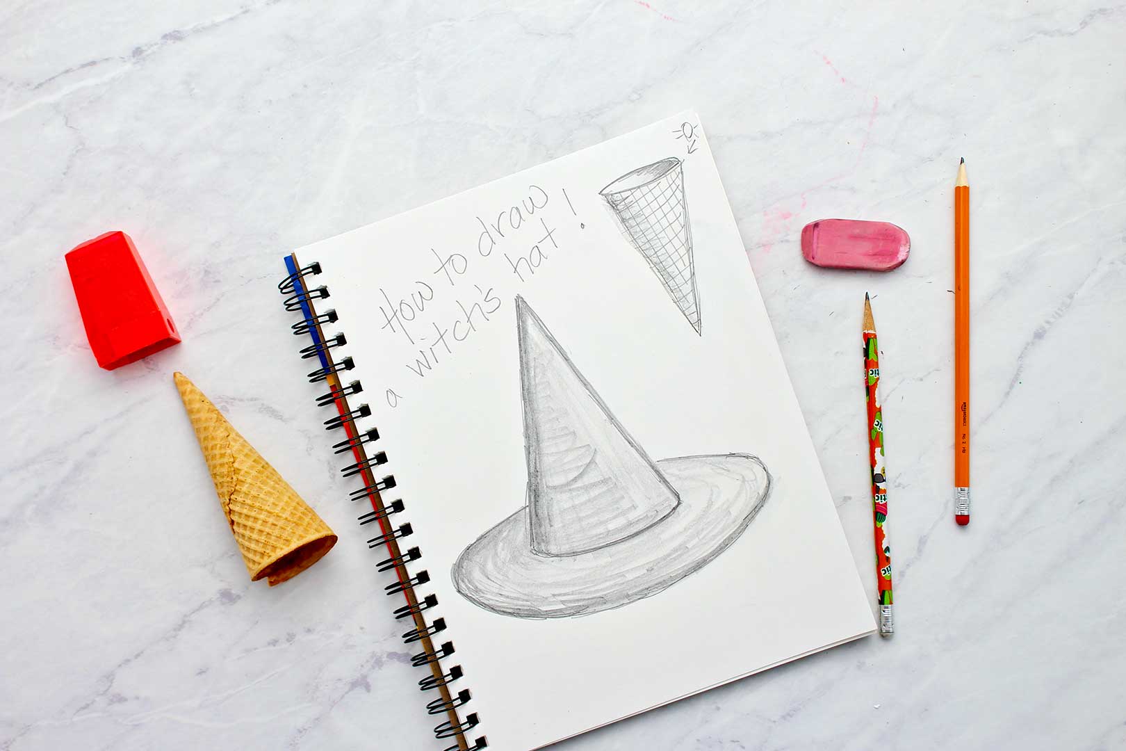 Completed sketch book page of witches hat and ice cream cone with pencils, pencil sharpener, eraser and ice cream cone resting near by.