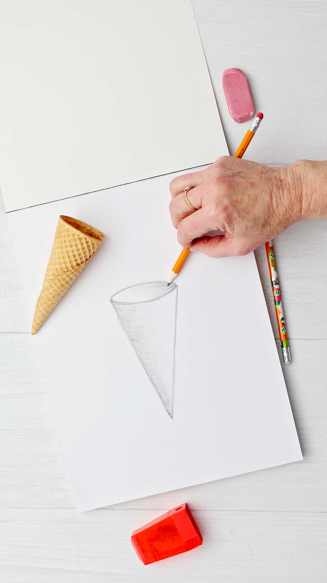 Hand shading the a sketch of a cone with an ice cream cone resting on paper.