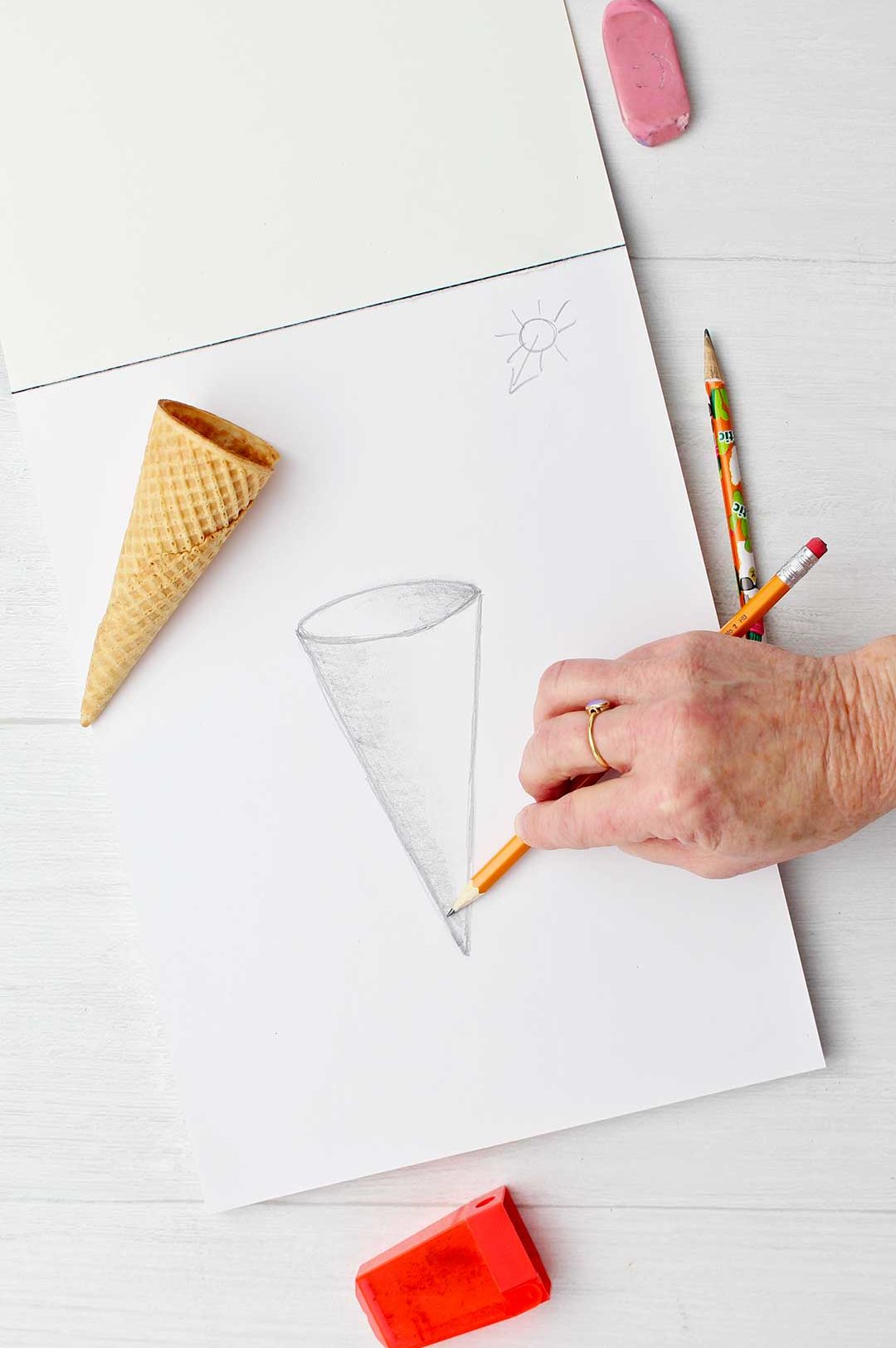 Hand shading the a sketch of a cone with an ice cream cone resting on paper.