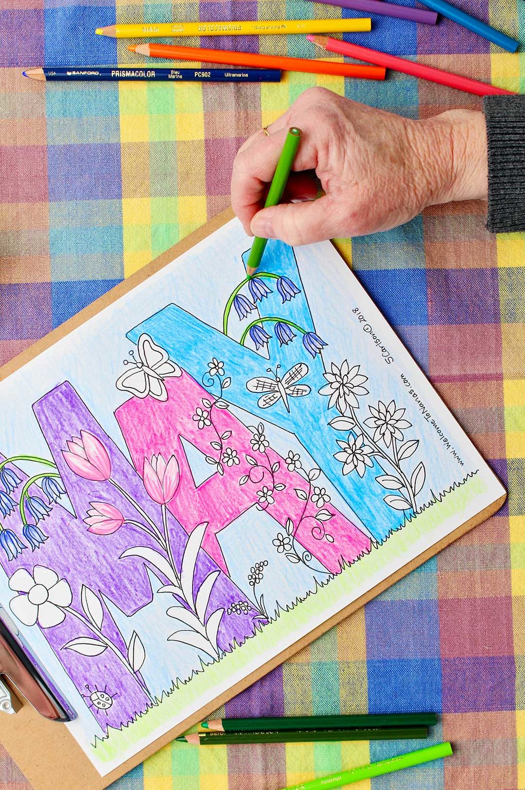 Hand coloring flower stem green in May Coloring Page with colored pencils resting on a colorful plaid background.