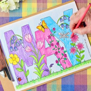 Hand coloring flowers pink in an almost completed May Coloring Page with colored pencils resting on a colorful plaid background.