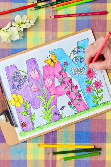 Hand coloring flowers pink in an almost completed May Coloring Page with colored pencils resting on a colorful plaid background.