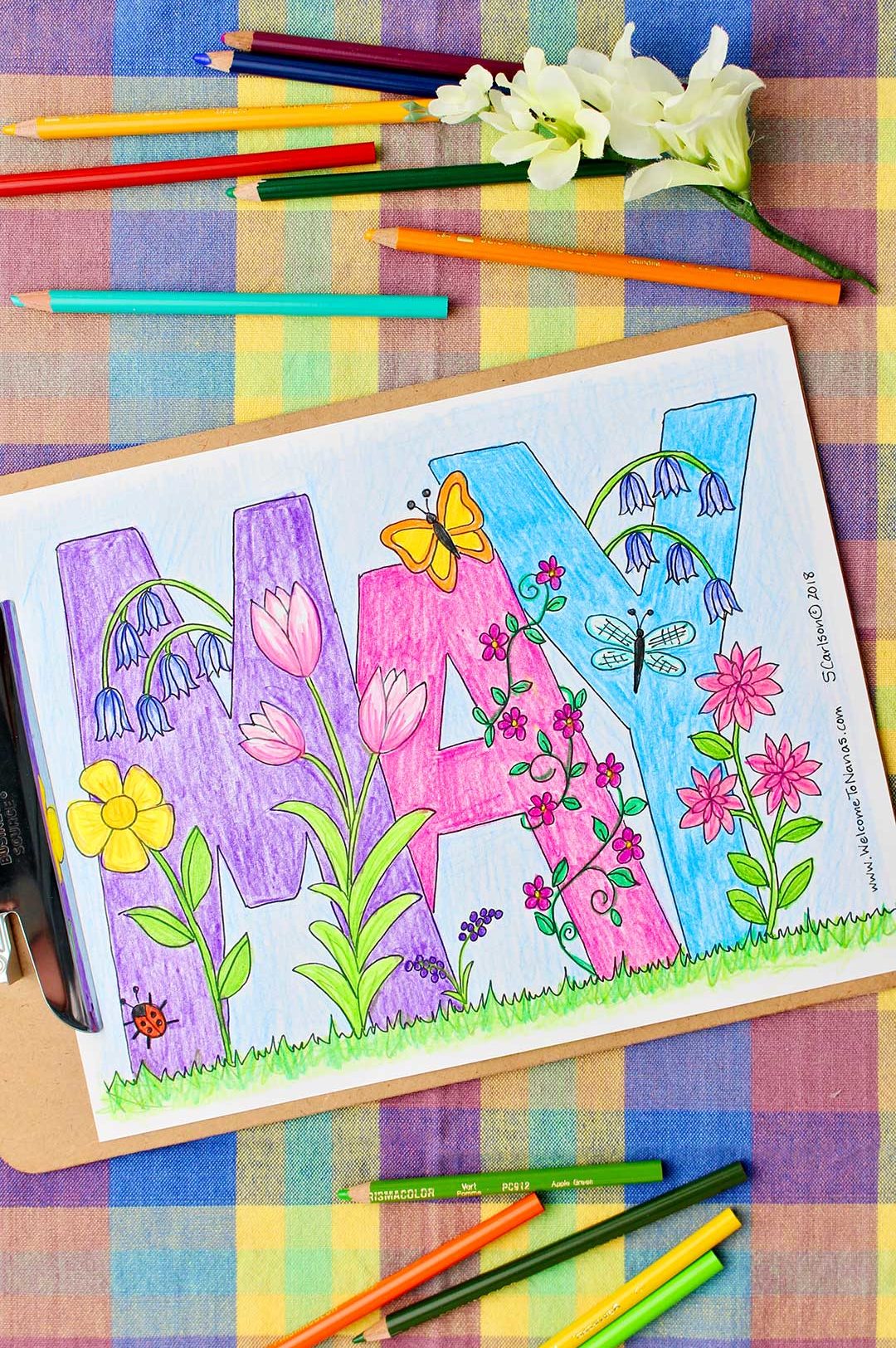 Completed May Coloring Page on a clipboard and colored pencils and flower resting on colorful plaid background.