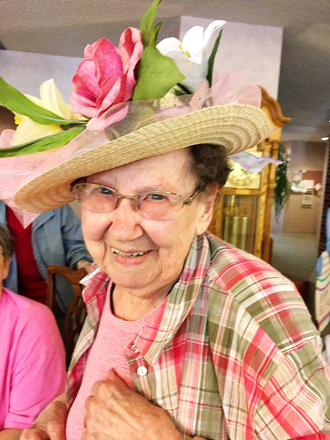 Photo of a beautiful older woman wearing a straw hat with flowers on it and a plaid green and pink shirt.