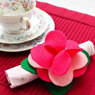 Completed felt flower napkin ring made from green and pink felt holds a rolled up napkin resting on a dark pink placemat with tea cup and saucer near.