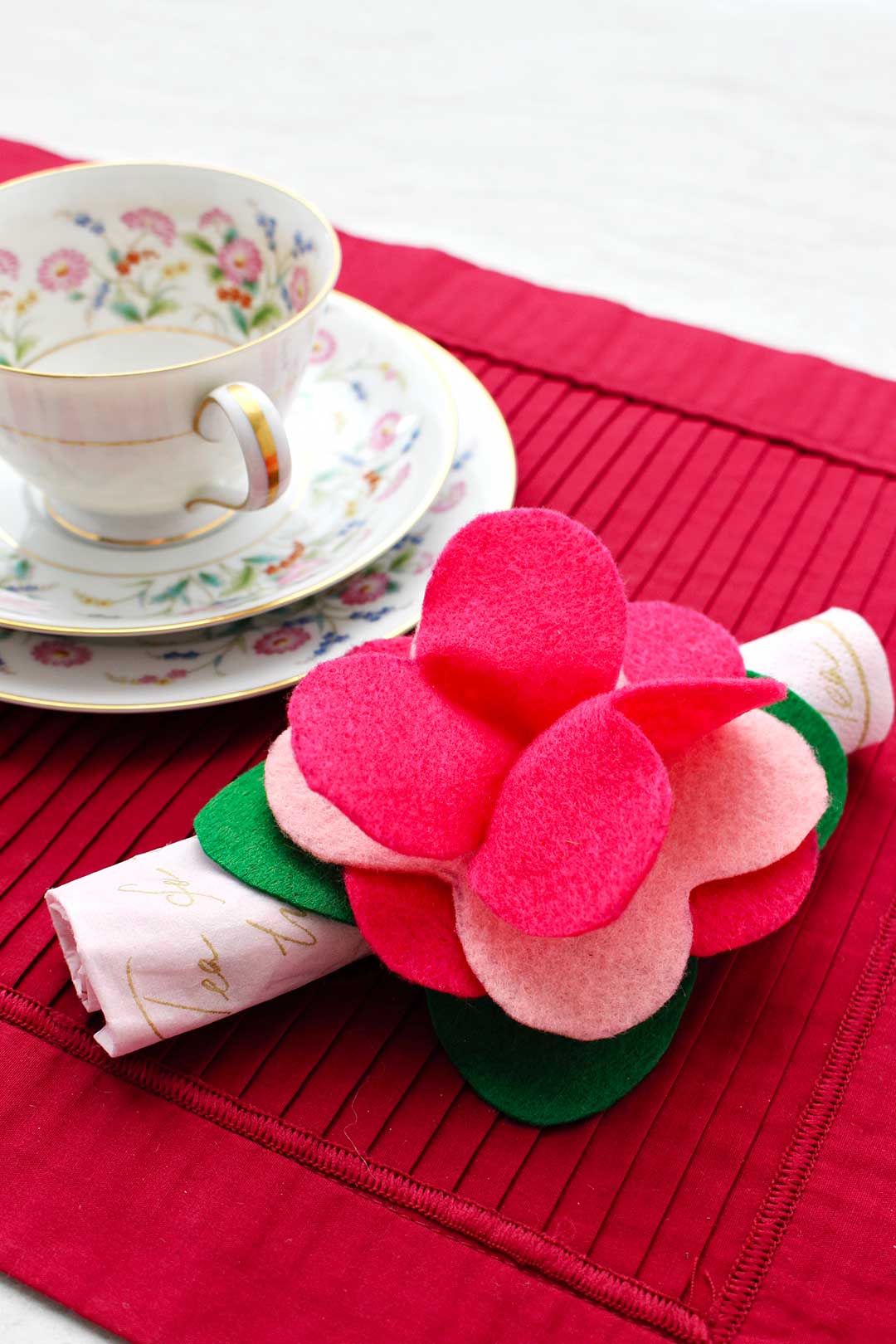 Completed felt flower napkin ring made from green and pink felt holds a rolled up napkin resting on a dark pink placemat with tea cup and saucer near.