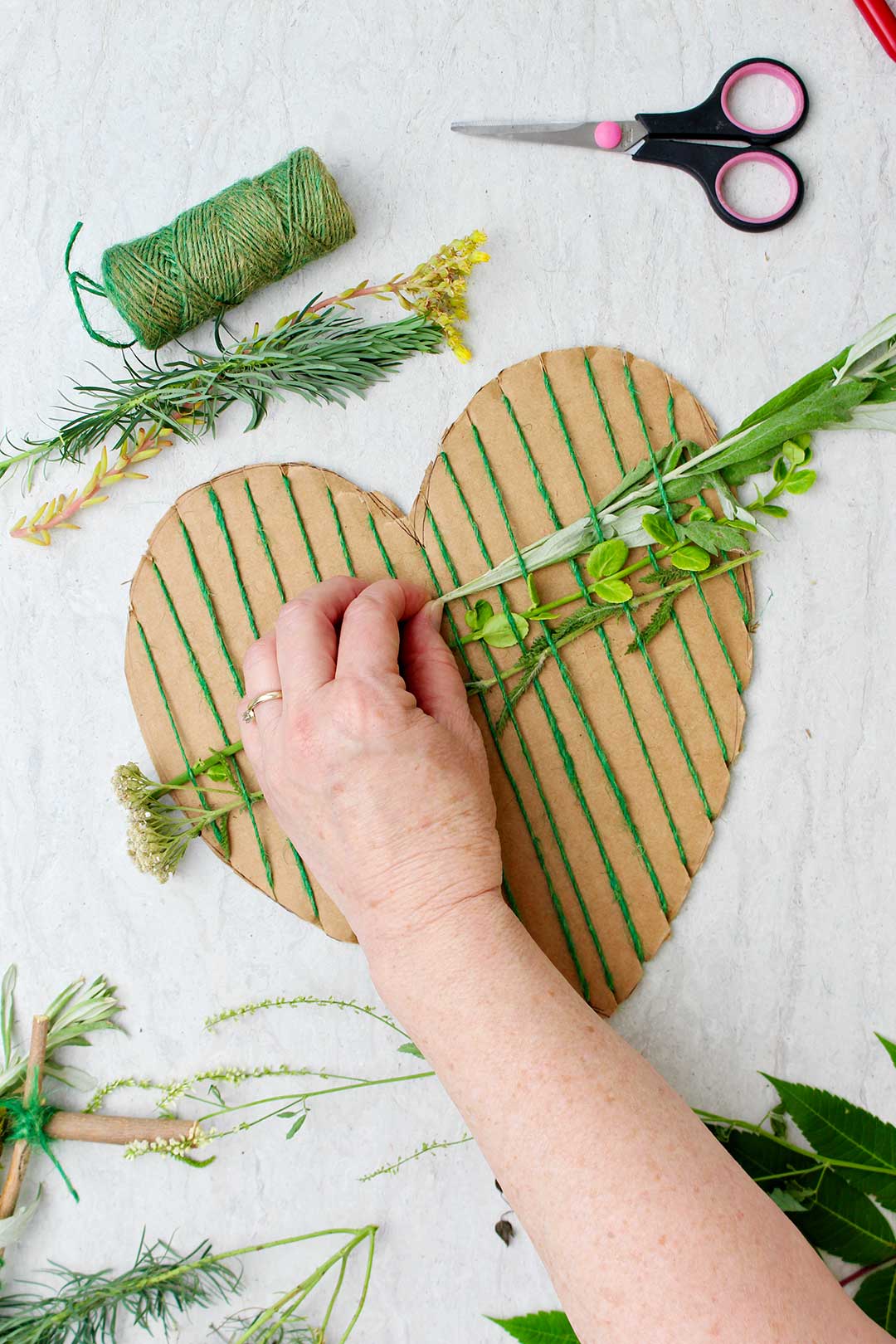 Hand weaving in pieces of greenery on heart shaped cardboard weaving project with supplies scattered near by.