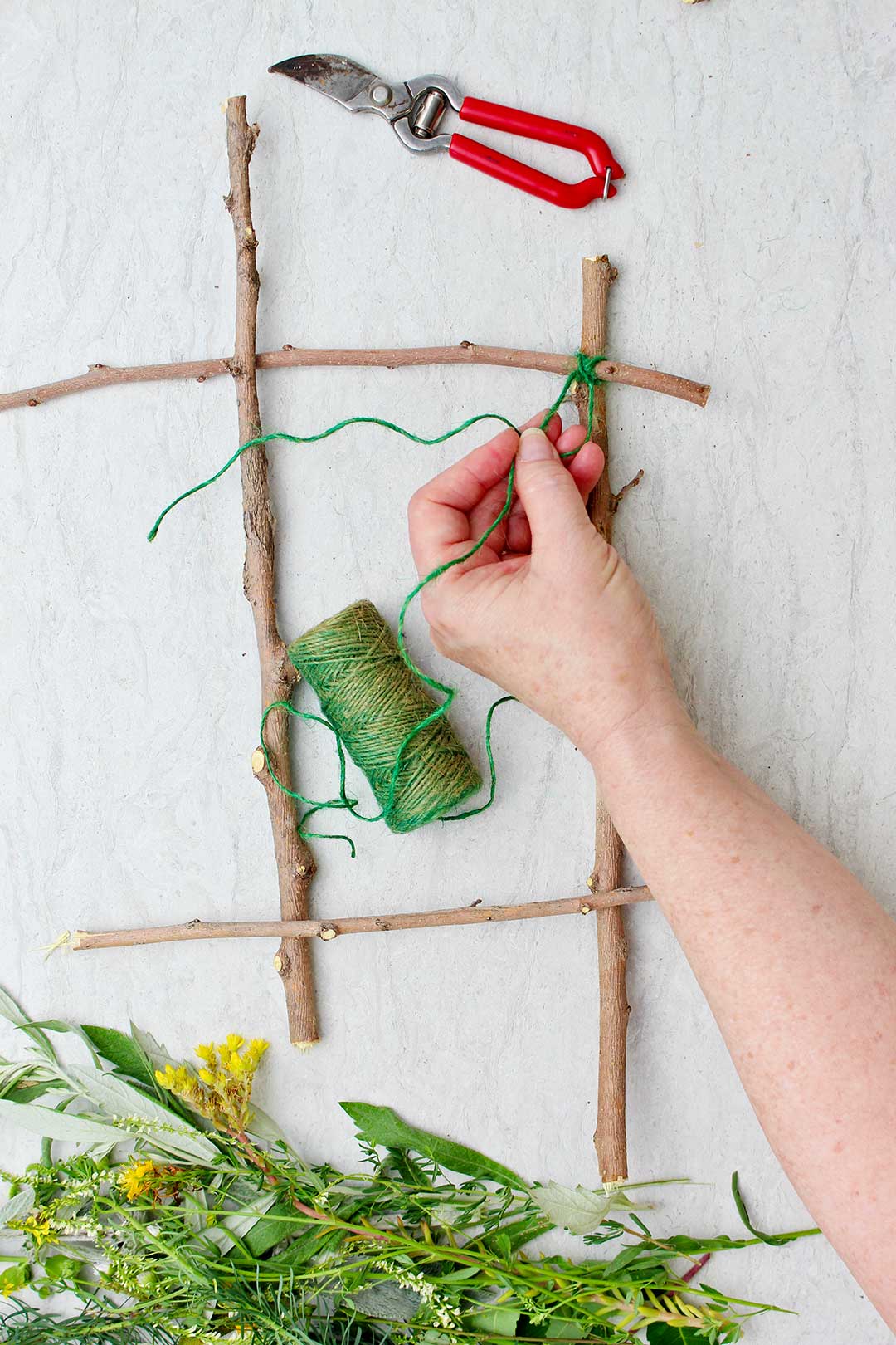 Hand securing green string to the crosspoint of two twigs on nature weaving project.