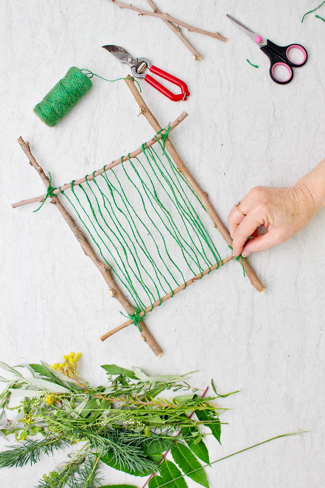 Hand wrapping green string around loom made from twigs with supplies for nature weaving near by.
