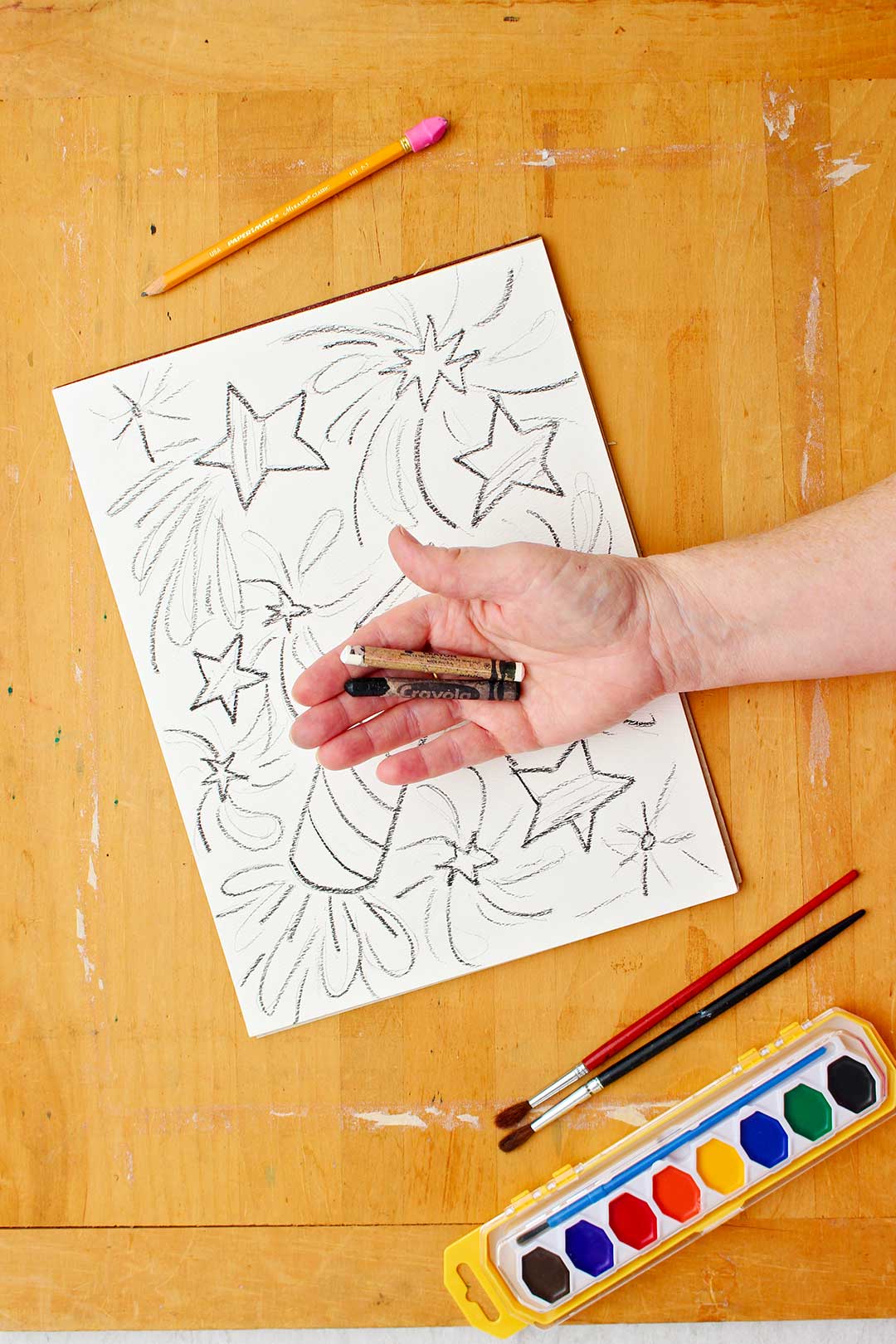 Hand holding a white and black crayon above a drawing of stars and rockets.
