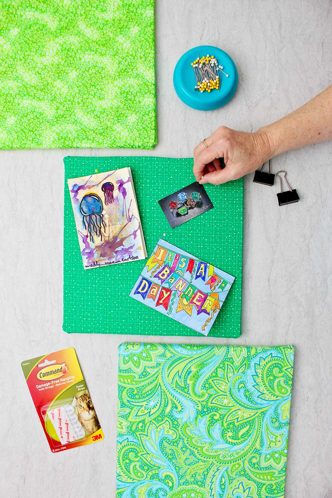 Hand pinning a photo to a foam core fabric bulletin board in a kelly green fabric with the other two completed boards near by.