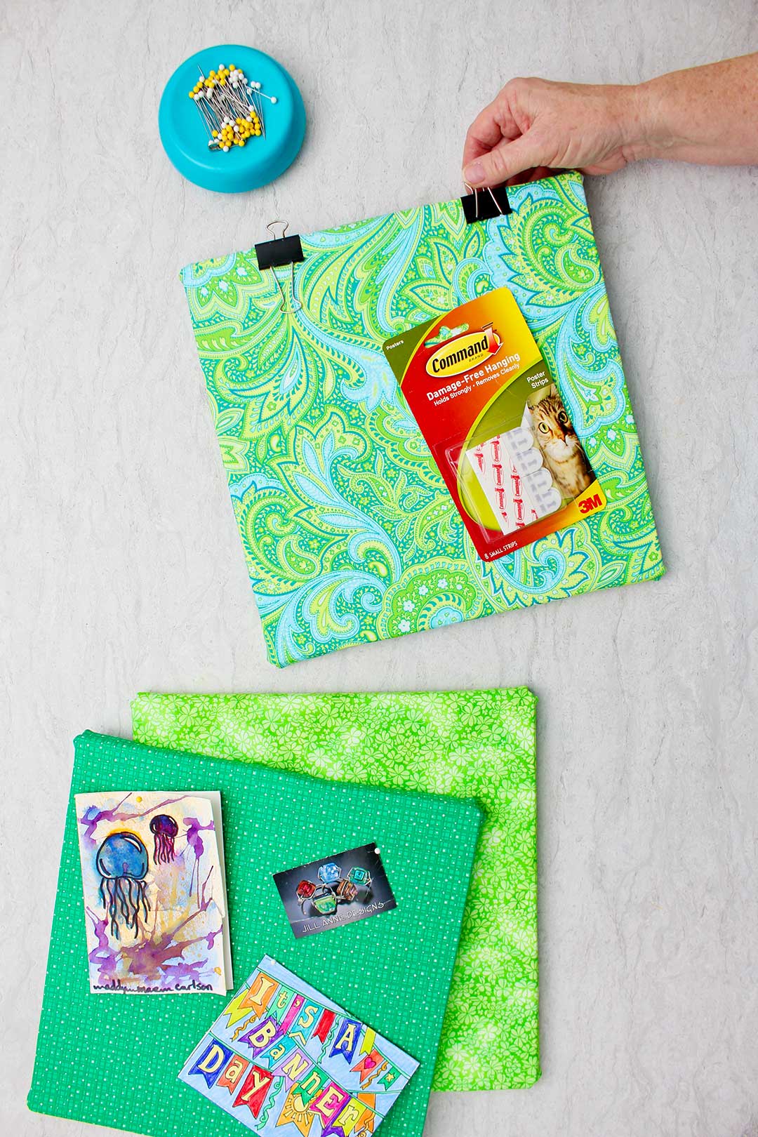 Hand securing binder clip the top of a foam core bulletin board in green paisley fabric.