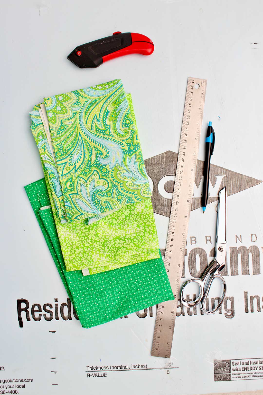 Supplies for fabric bulletin boards with foam core. Foam core, three different patterned green fabric, box cutter, pen, scissors and ruler.