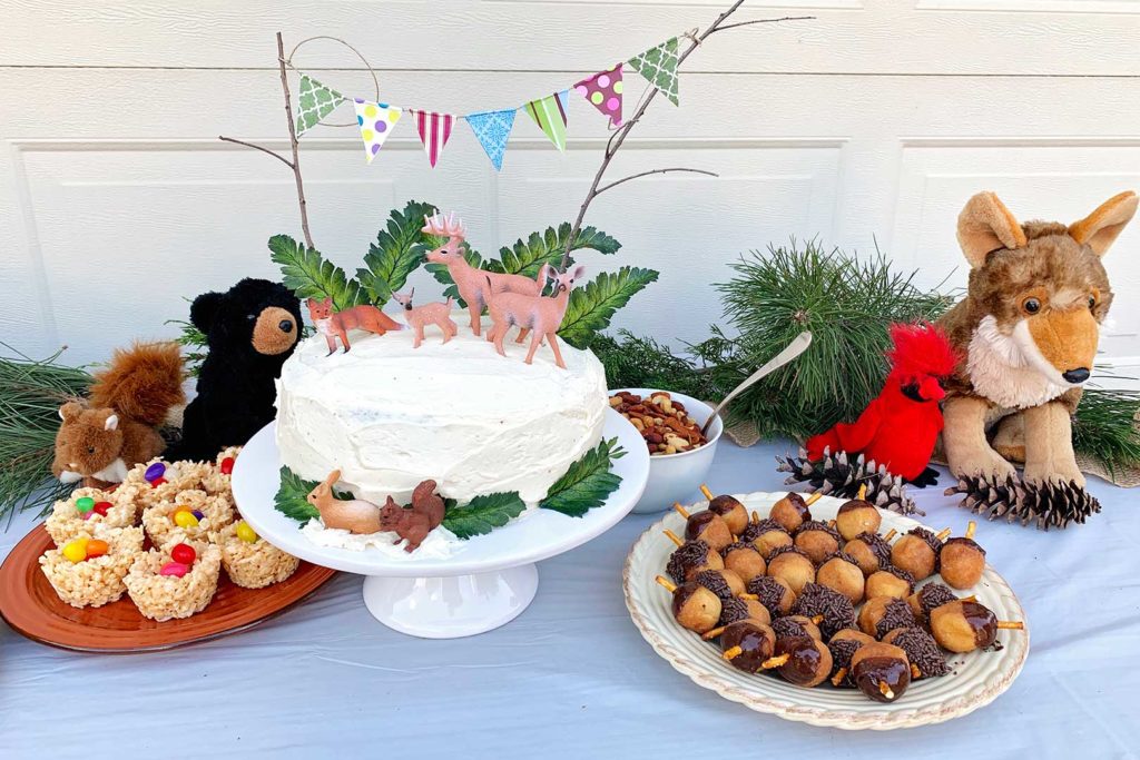 Woodland party table decor. A cake with toy deer, greenery, acorn donuts, stuffed animals and rice crispy candy nests.
