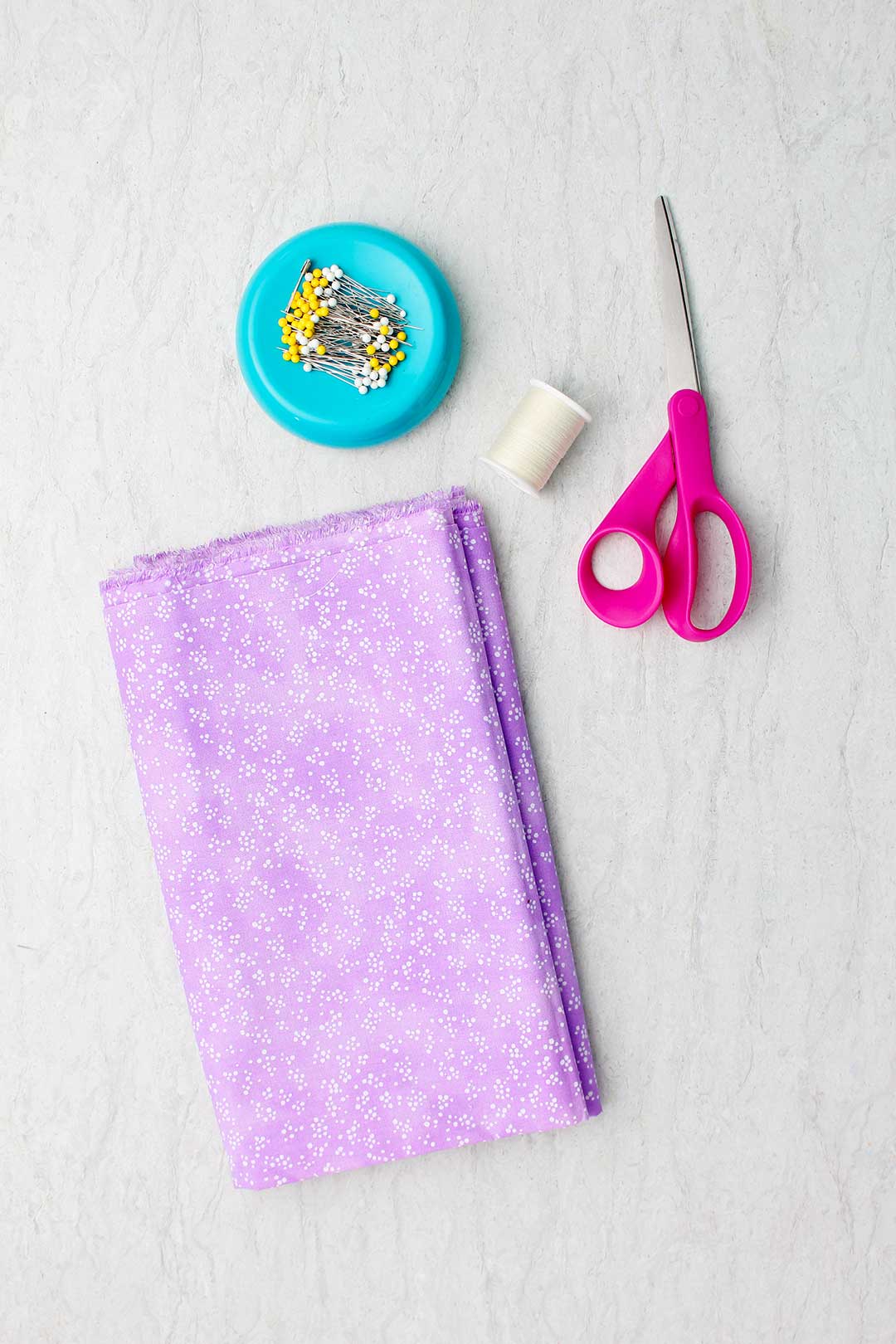 Purple fabric with white dots, white thread, pink scissors and a blue magnetic straight pin holder with pins.