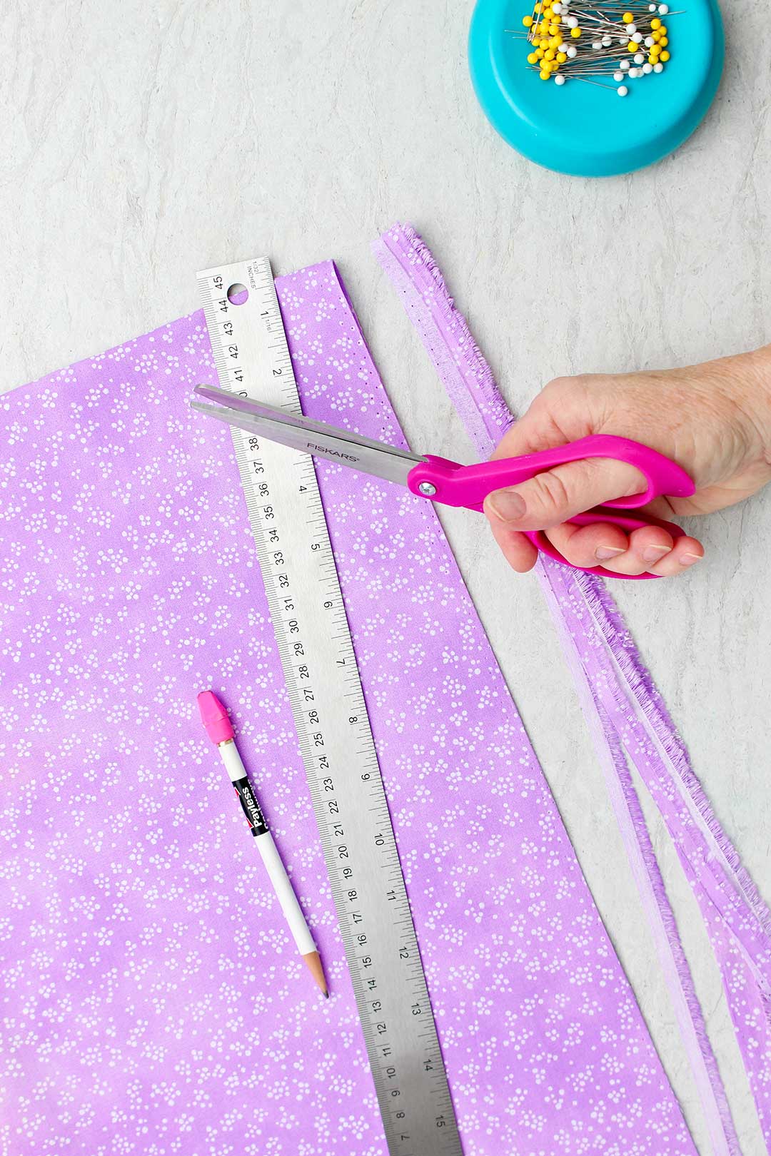 Hand holding a pair of pink scissors near purple fabric with the edge cut off and a metal ruler, straight pin holder and pencil.