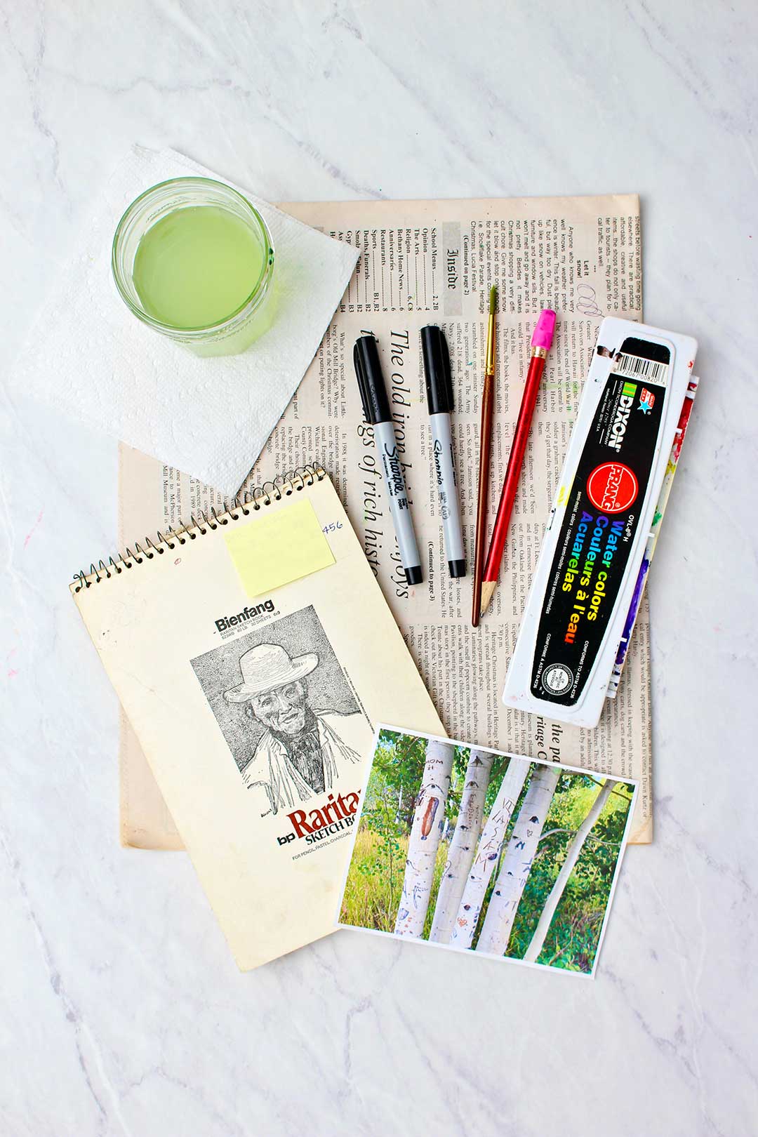Supplies for aspen tree watercolor painting. Sketch pad, watercolors, inspiration photo, cup of water, newsprint and a paper towel.