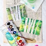 Vertical view of completed aspen water color painting resting on a piece of newspaper with inspiration photo, watercolors, markers, paintbrush and cup of water near by.