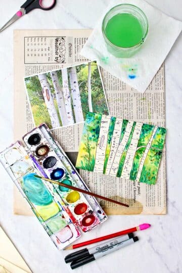 Vertical view of completed aspen water color painting resting on a piece of newspaper with inspiration photo, watercolors, markers, paintbrush and cup of water near by.