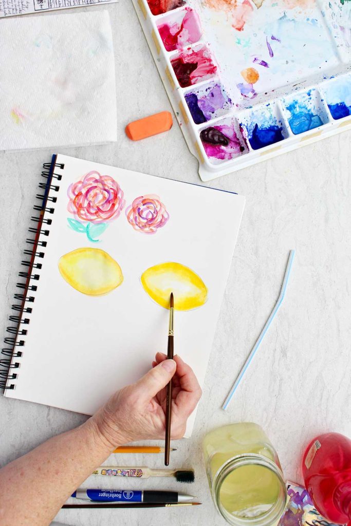 Hand painting a lemon with watercolor supplies near by.