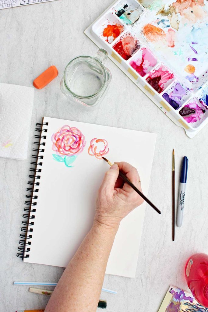 Hand painting flowers on a piece of white paper with watercolor supplies near by.