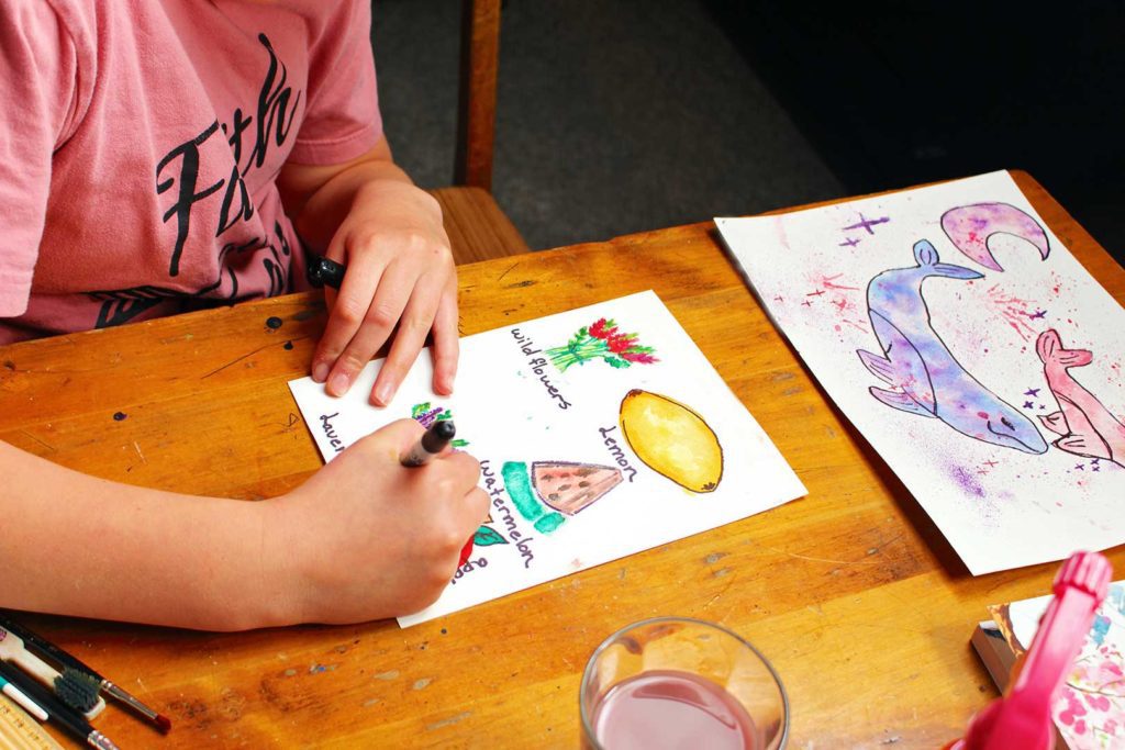 Young girl labeling some watercolor images she painted at a kitchen table.