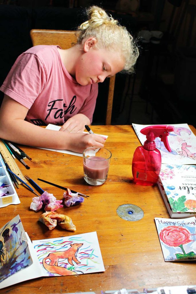 Young girl with blonde hair and a pink shirt sits at a kitchen table with many paintings using watercolors around her.