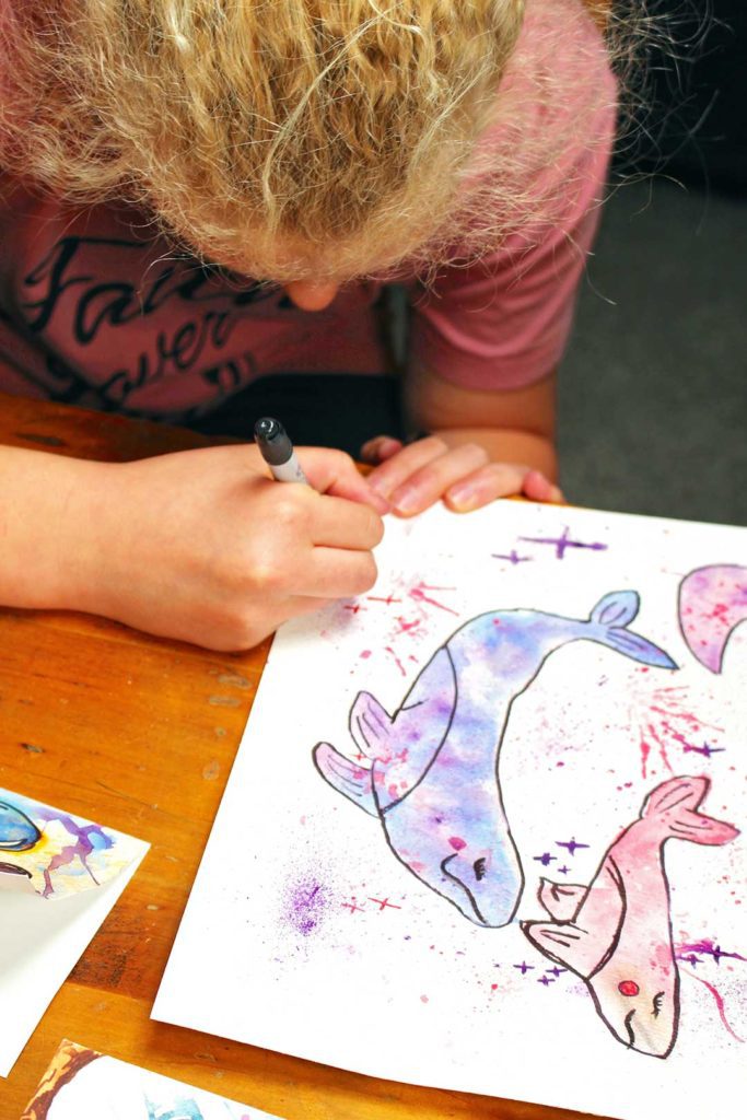 Young girl with blonde hair and a pink shirt drawing details on her watercolor of some whales.