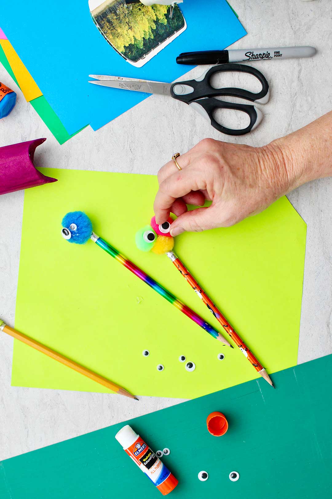 Hand placing google eyes on pom poms on top of a pencil.