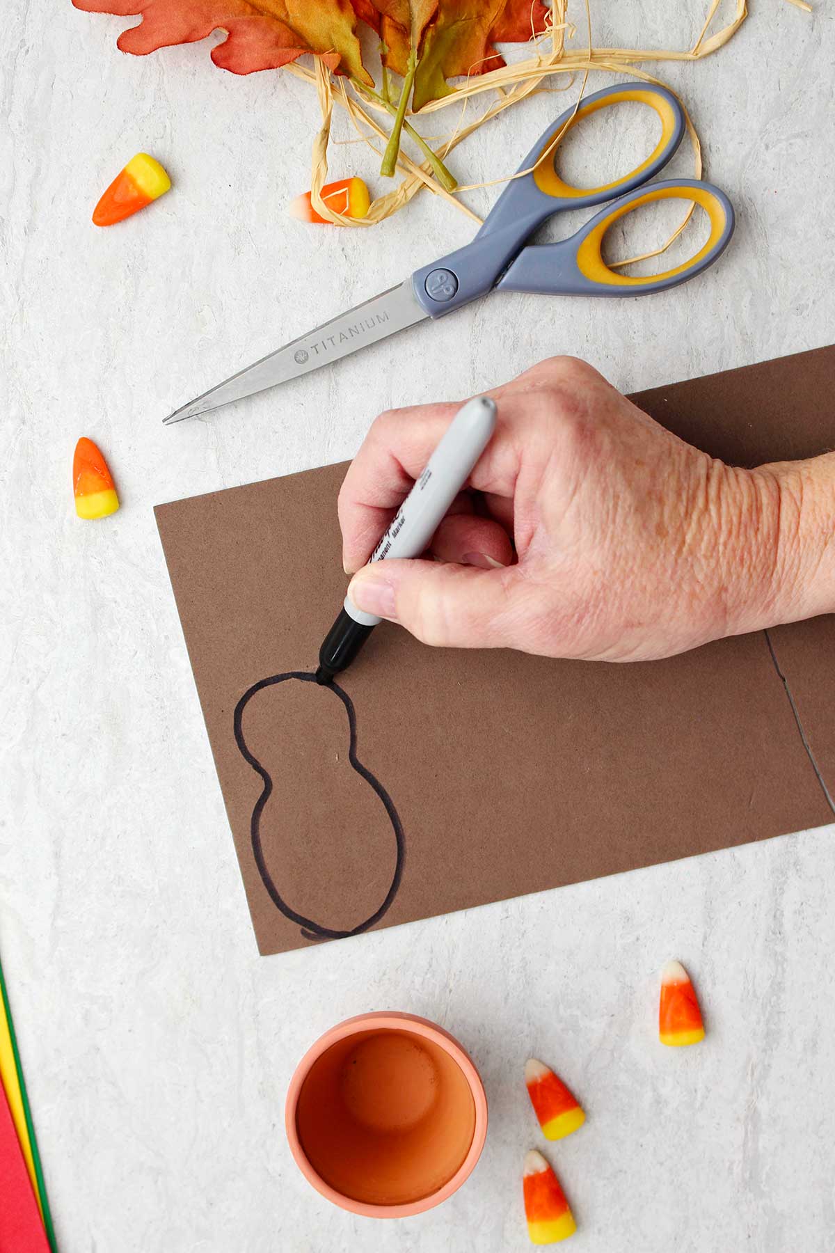 Hand using sharpie to draw a peanut shape out of brown paper for the turkey body with supplies near by.