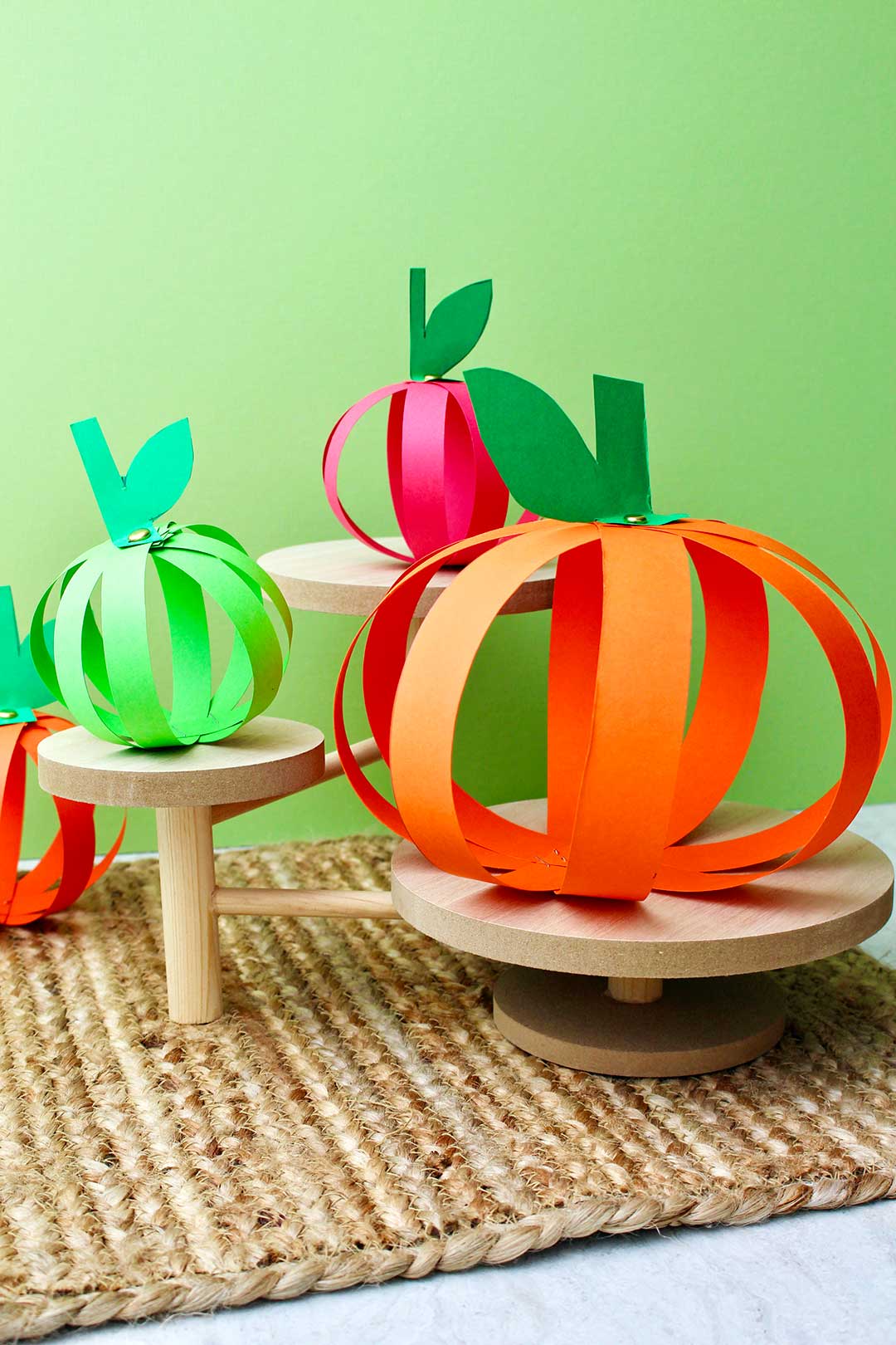 Vertical image of four completed paper strip pumpkins sitting on wooden pedestals against a green background.