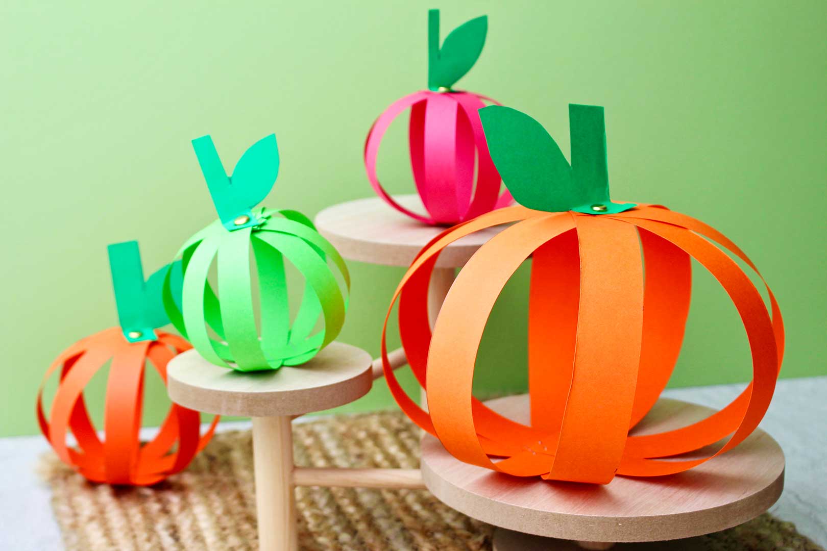Four completed paper strip pumpkins sitting on wooden pedestals against a green background.