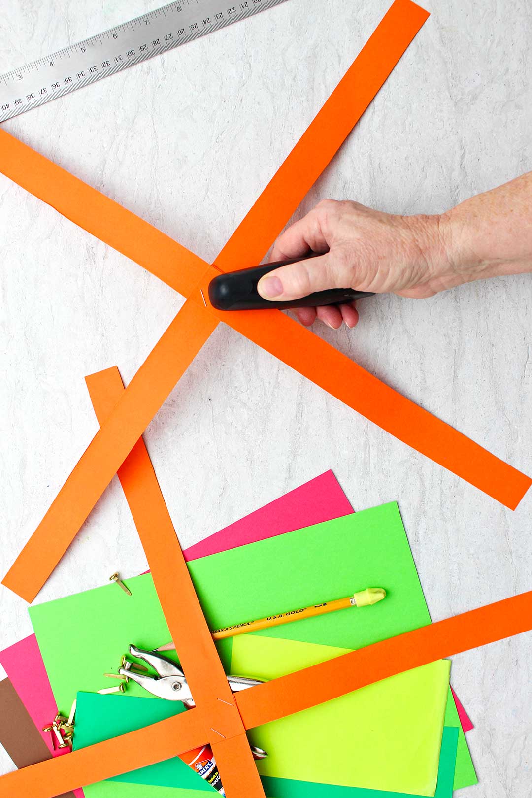 Hand stapling four strips of orange paper into "x" shape.