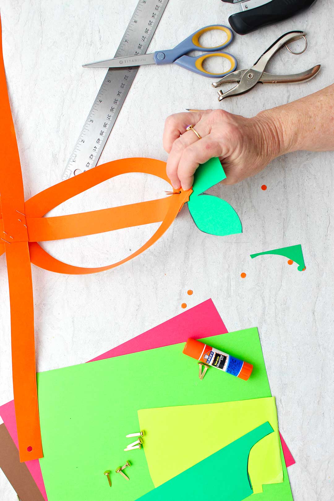 Hand securing green paper stem with metal brad on partially made orange paper pumpkin.