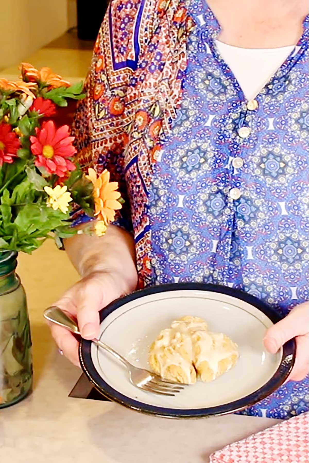 Woman in blue and orange print top holds a plate with an iced pumpkin scone near some flowers in a mason jar.