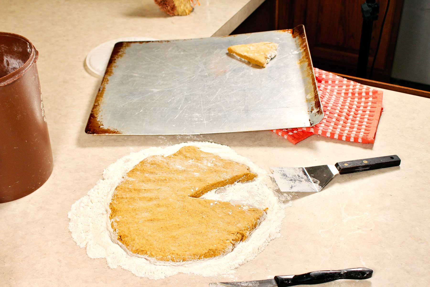Circular shaped dough with pie shaped piece missing with tray and scone on it in the background.
