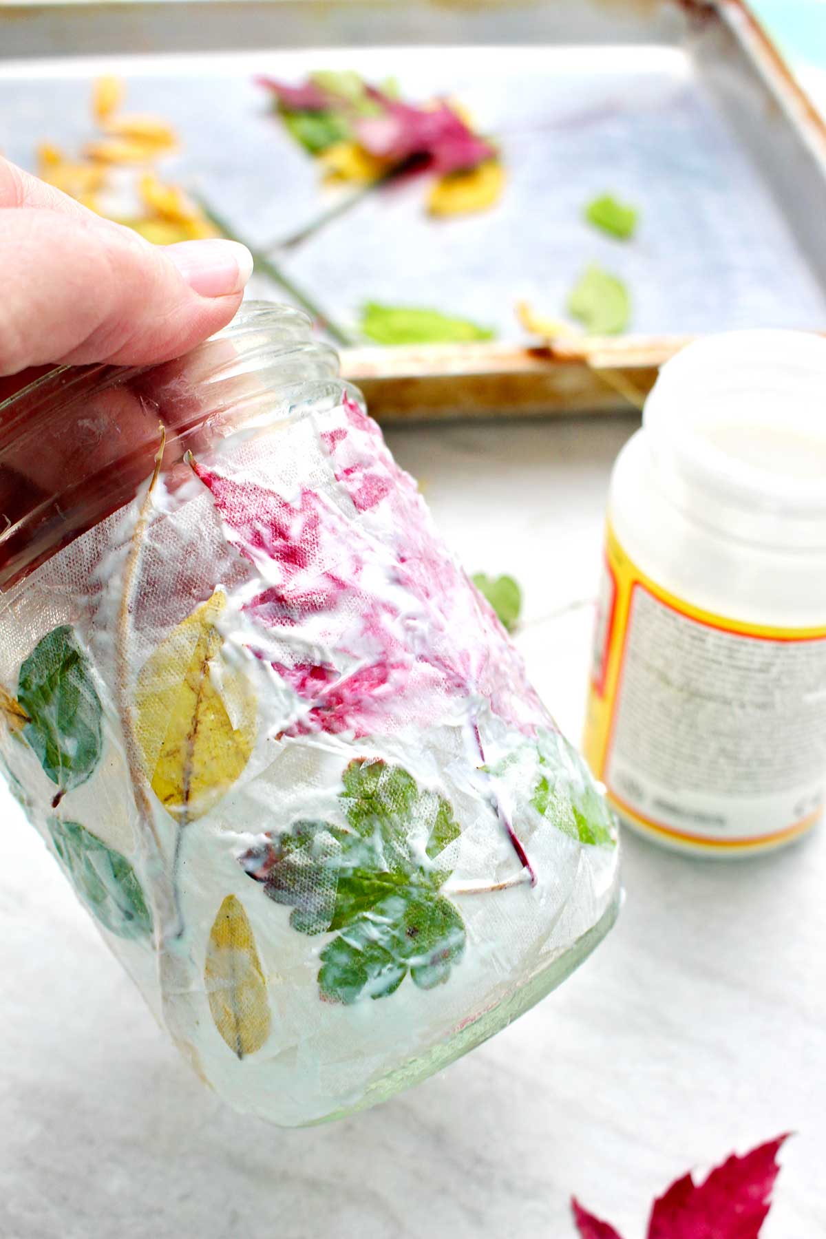 Close up view of hand holding jar with drying Mod Podge, paper towel and leaves on a mason jar.