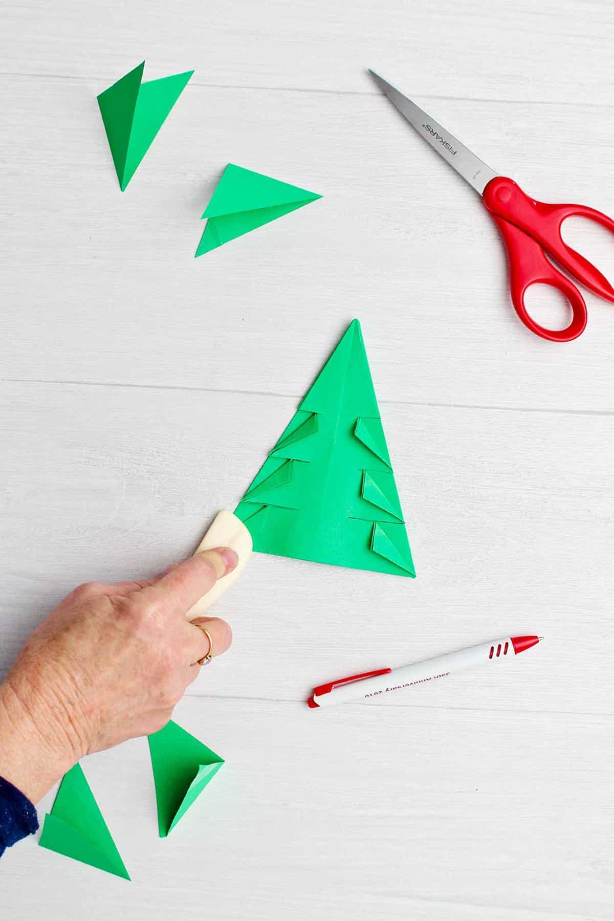 Hand using paper creaser to cleanly fold down pieces of green origami tree.