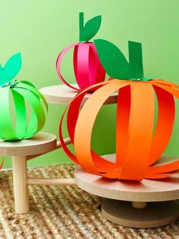 Four completed paper strip pumpkins sitting on wooden pedestals against a green background.