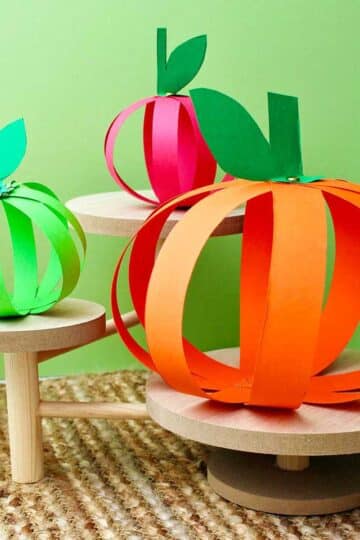 Four completed paper strip pumpkins sitting on wooden pedestals against a green background.