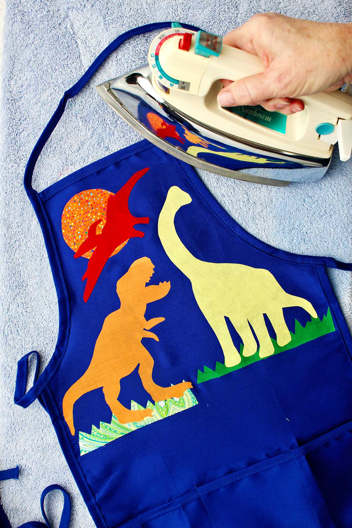 Hand holding iron over almost complete dinosaur decorated blue apron.