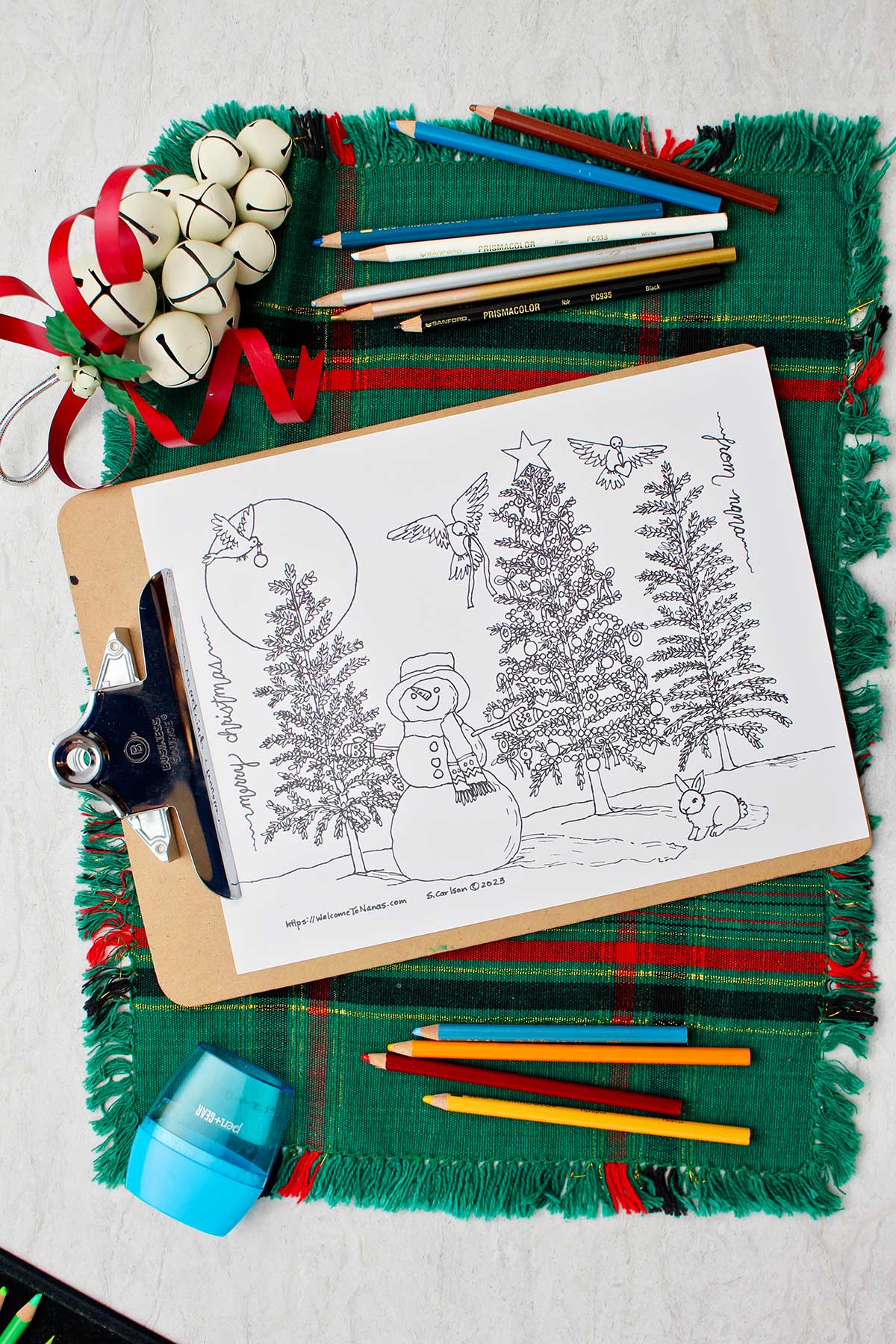 Uncolored Christmas tree coloring page on a clip board resting on a green and red placemat with colored pencils, a pencil sharpener and bells near by.