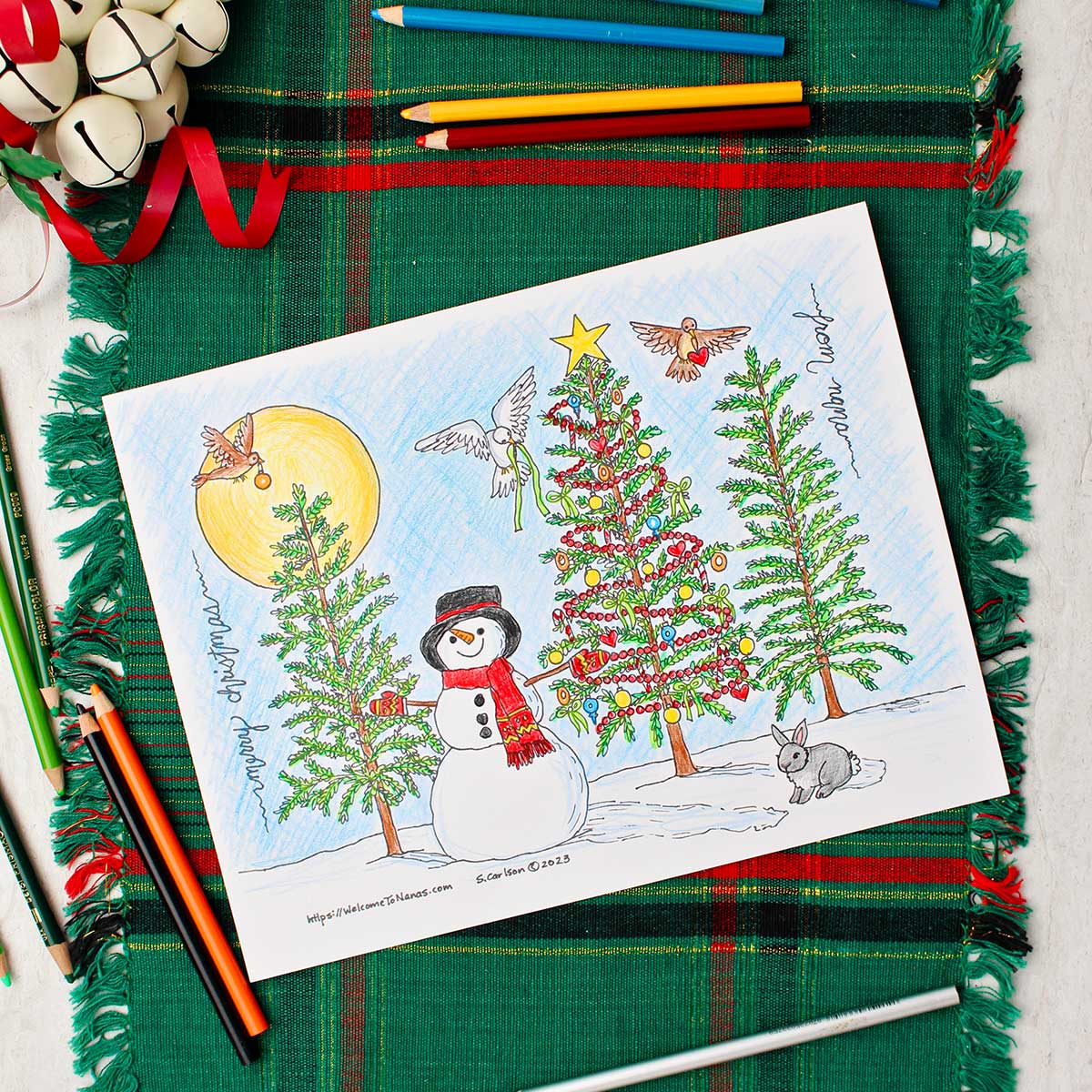 Completed Christmas tree coloring page resting on a green and red placemat with white decorative bells near by. Square image.