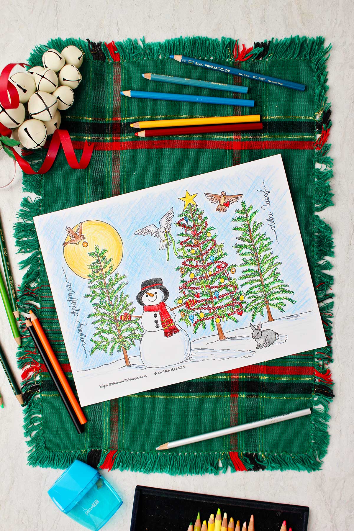 Completed Christmas tree coloring page resting on a green and red placemat with white decorative bells and colored pencils near by.