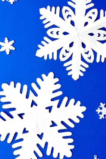 Two completed snowflakes made from white paper resting on blue background.